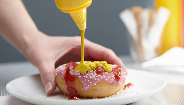 A woman's hand putting mustard on a donut that's already covered with ketchup