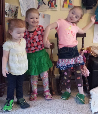 Three little girls wearing colorful and silly clothes are smiling together in a room filled with dra...