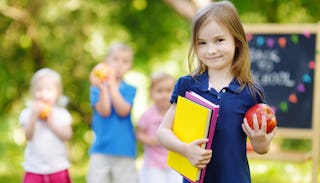 Little girl ready for kindergarten holding one apple and three books in her arms