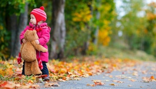 A little girl in pink clothes holding her big teddy bear on the forest road in the autumn