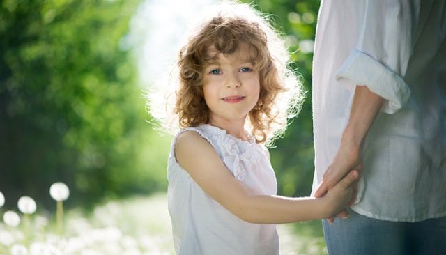 A smiling curly-haired girl in a white top holding her mother's hand