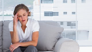 A stressed-out blonde woman with ADD in a white sleeveless shirt and gray pants sitting on a gray co...