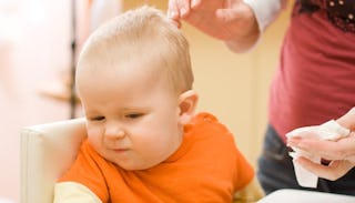 A toddler in an orange shirt turning away from the hands of his mother who is trying to wipe his fac...