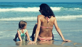 A brunette woman in a white and red striped bikini next to a baby in a green-white bathing suit sitt...