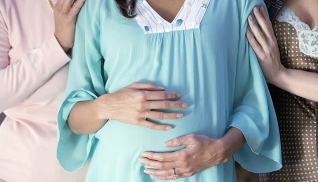 Two women holding the first-time mom while she is holding her pregnant belly in green pajamas who ha...