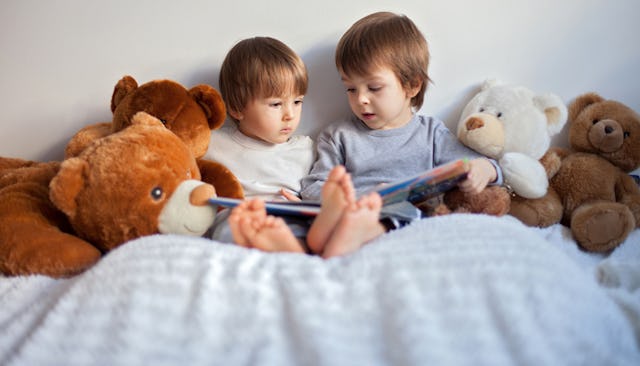 Two brown haired boys wearing a gray and white shirt, looking at a book with stuffed bear toys surro...
