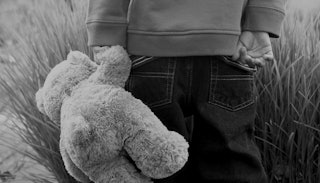 A small kid standing alone with a teddy bear in black and white