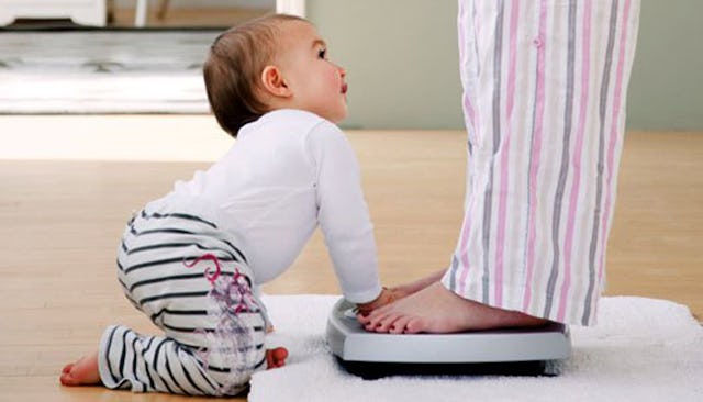 Woman on a scale with her baby leaning on it while sitting next to the scale