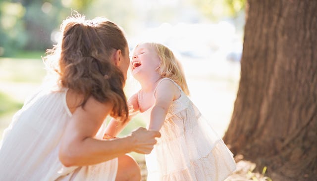 A brunette woman in a white dress holding her laughing four-year-old daughter next to a tree