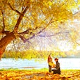 Mother and daughter playing near a lake under a tree with yellow leaves whose leaves are falling in ...