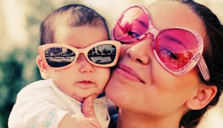 A smiling brunette 'sorry-not-sorry' mom with pink sunglasses holding a baby with oversized sunglass...