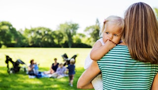A suburban mom in a green-white shirt holding her baby and other mothers blurred in the background