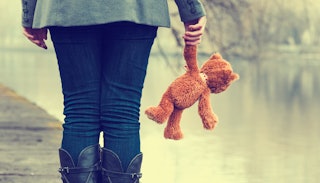 Woman standing with a teddy bear next to a river as she experiences the void as she's done having ch...