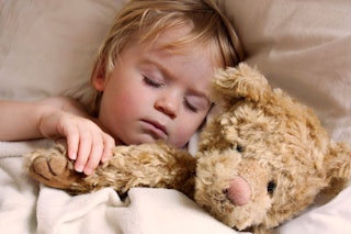 A little blonde boy sleeping in beige bed sheets holding his light brown teddy-bear