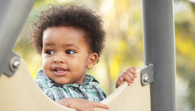 An adopted curly-haired toddler in a blue-green checkered shirt smiling at a playground