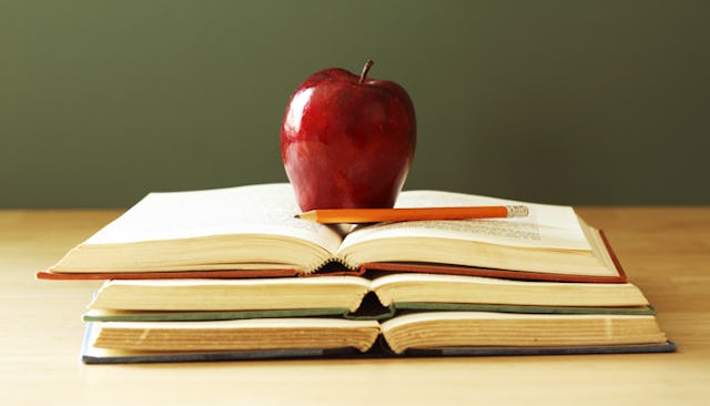 A red apple and an orange pencil on top of a stack of three books on a wooden surface and a sage gre...