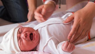 A newborn baby crying in a white onesie while parents are standing next to it