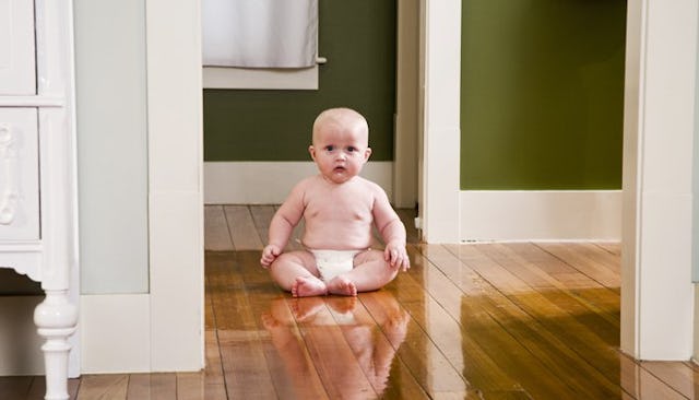 A baby with a neutral facial expression in a diaper sitting on a wooden floor