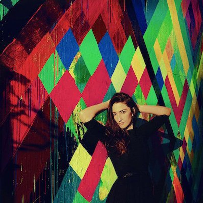 Sarah Baraz in a black dress with her hands behind her head in front of a multi-colored wall