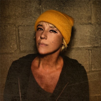 Cat Power in front of a brick wall wearing a black jacket and yellow knit beanie