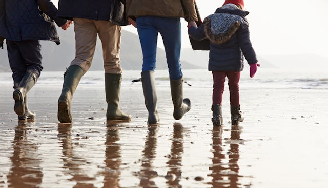Two adults and two children holding hands and walking in a row on a beach wearing dark green rubber ...