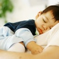 A sleep-deprived dad and a dark-haired toddler in a dark blue shirt and light blue pants sleeping on...