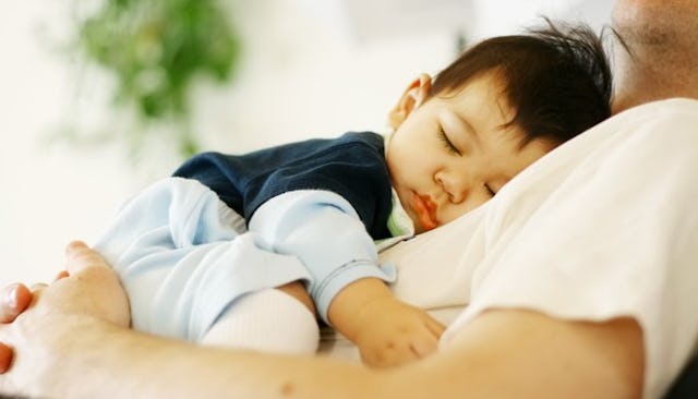 A sleep-deprived dad and a dark-haired toddler in a dark blue shirt and light blue pants sleeping on...