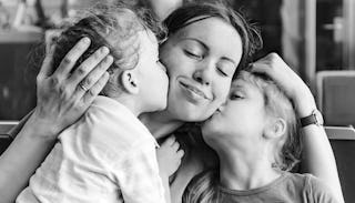 A mom with closed eyes, smiling and hugging her kids while they're giving her kisses on her cheeks i...