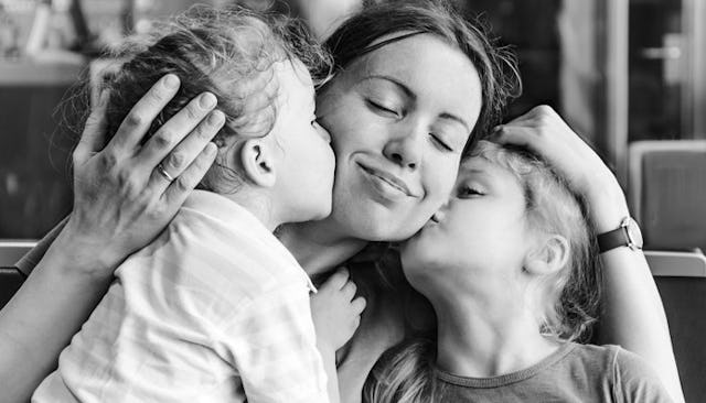 A mom with closed eyes, smiling and hugging her kids while they're giving her kisses on her cheeks i...
