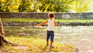 A toddler boy in a white shirt and blue denim jeans standing next to a river with his back turned