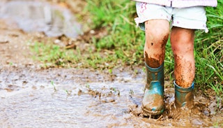 A little girl playing in a mud puddle in little green rain boots
