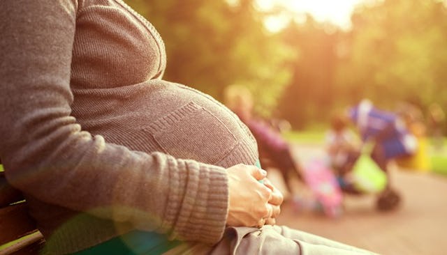 A gestational surrogate sitting on the bench while holding her stomach in a sunny park