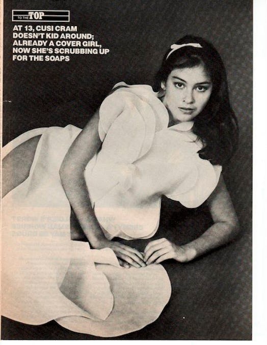 Cusi Cram posing on the floor, in a white flowy dress, with a white headband in her luscious brown h...