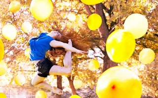 A red-haired woman surrounded by yellow balloons jumping enthusiastically, with her foot almost touc...