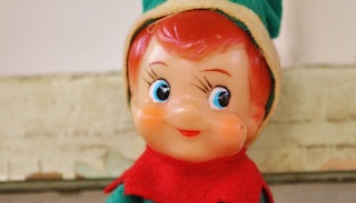 A red-haired elf on the shelf with blue eyes, red hair, and a green hat