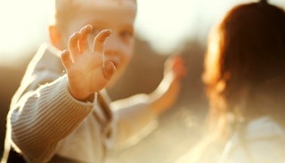 A boy with Autism in a beige knit sweater waving towards the camera with his mother blurred in the b...