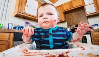 A baby boy making a mess with food while sitting in his baby seat.