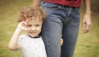 A toddler boy walking while holding his mother's right leg with his left hand