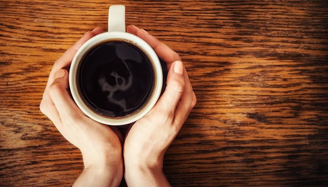 A woman holding a cup of coffee with both of her hands on a wooden surface