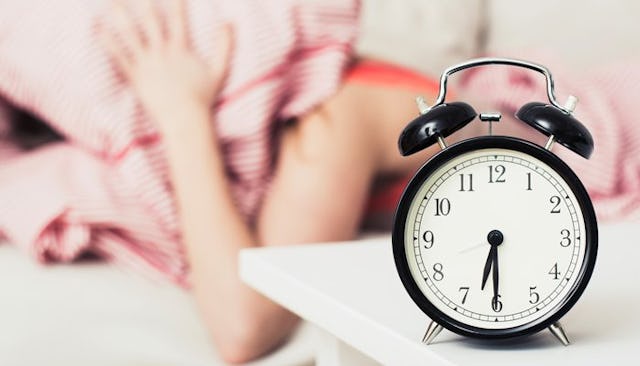 A close-up image of an alarm clock with an exhausted new mom in the background ignoring the alarm wi...