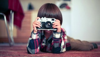A brown-haired child in a plaid shirt and brown pants lying down and looking through a camera lens