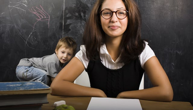 An elementary teacher smiling as she sits at a table in the classroom while a kid is playing behind ...