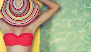 A woman in a red bikini and a hat over her head lying on an inflatable air bed in a pool who's not s...