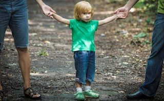 Blonde child wearing a green shirt and shoes, and blue pants holding hands of both of his parents wi...