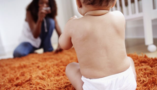A baby in a diaper sitting on a yellow carpet with the mother blurred in the distance, taking a pict...
