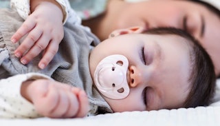 A baby peacefully sleeping with a pacifier in her mouth and his mom sleeping behind her