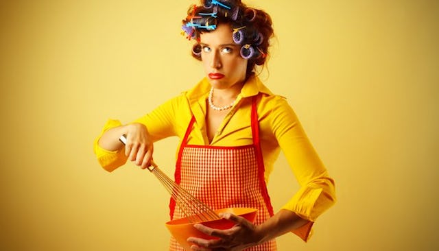 A housewife in a yellow shirt, red apron, and hair curlers mixing food in a bowl with a bored facial...