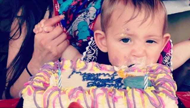 A brown haired boy being held by his mother, biting into a white cake