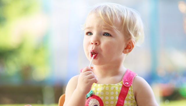 A young blonde child wearing a yellow and pink dress with a lollipop in her mouth