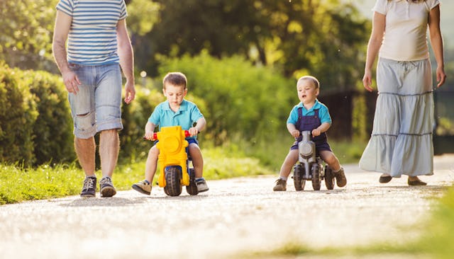 Two toddlers riding toy cars down a street with their mom and dad walking on both sides next to them